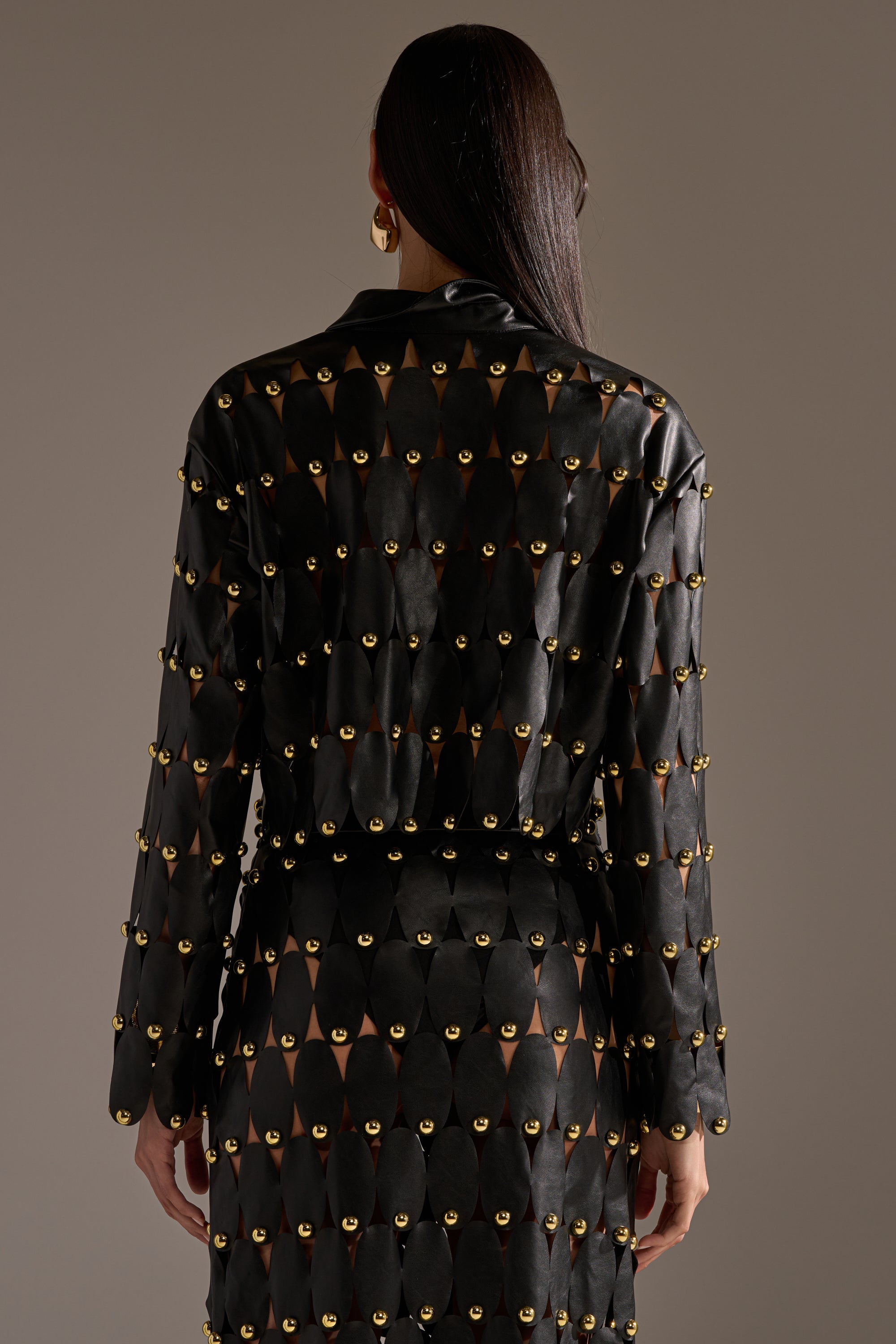 A person faces away, wearing THE SNEAK PEEK STUDDED PU BLOUSE IN BLACK, featuring gold metallic studs and triangular cutouts that reveal skin. Their straight dark hair cascades down their back, paired with gold hoop earrings. 5
