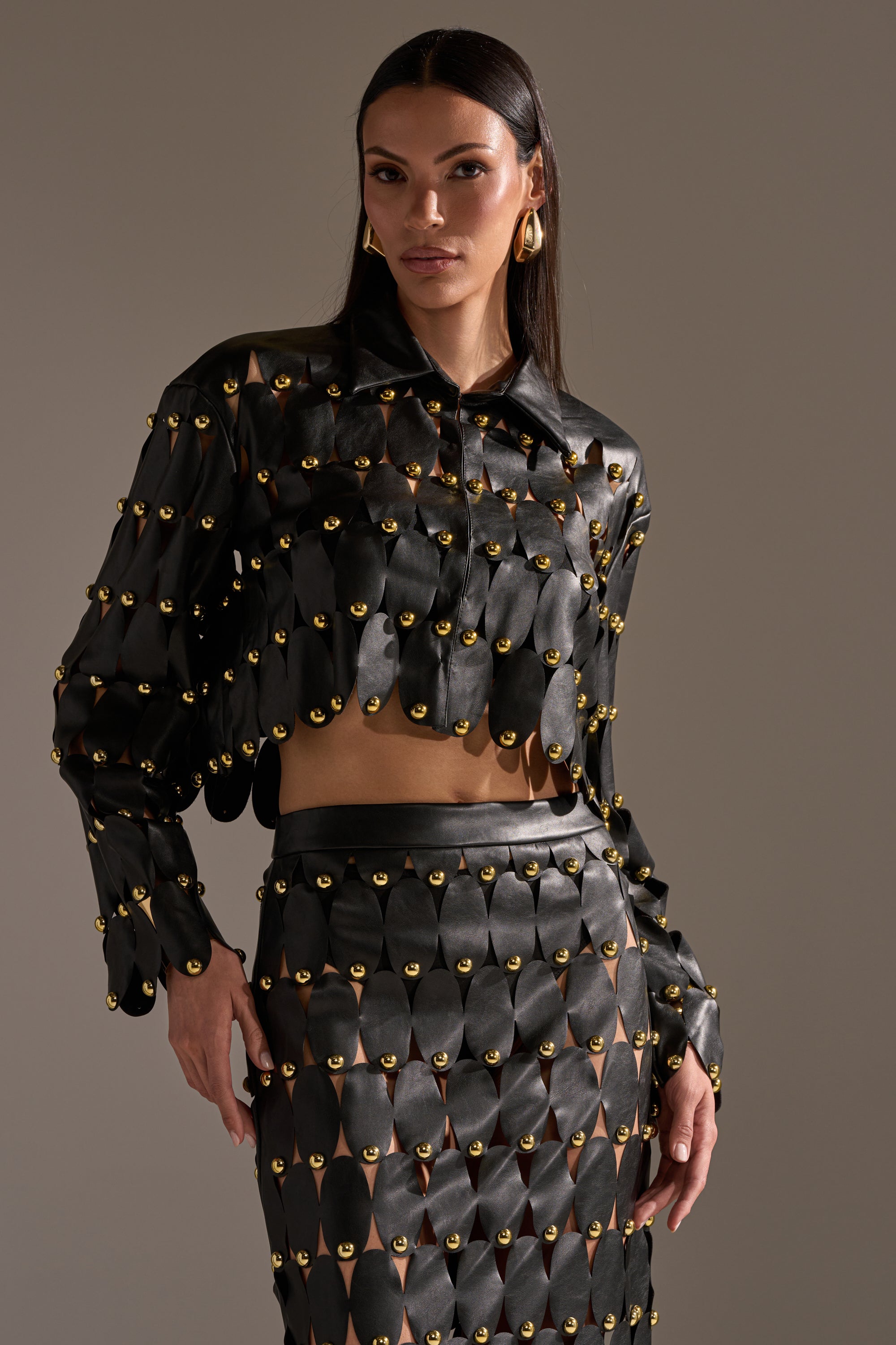 A woman wears THE SNEAK PEEK STUDDED PU BLOUSE IN BLACK with matching scalloped-panel skirt and gold grommet accents, posing against a plain, neutral background. 6