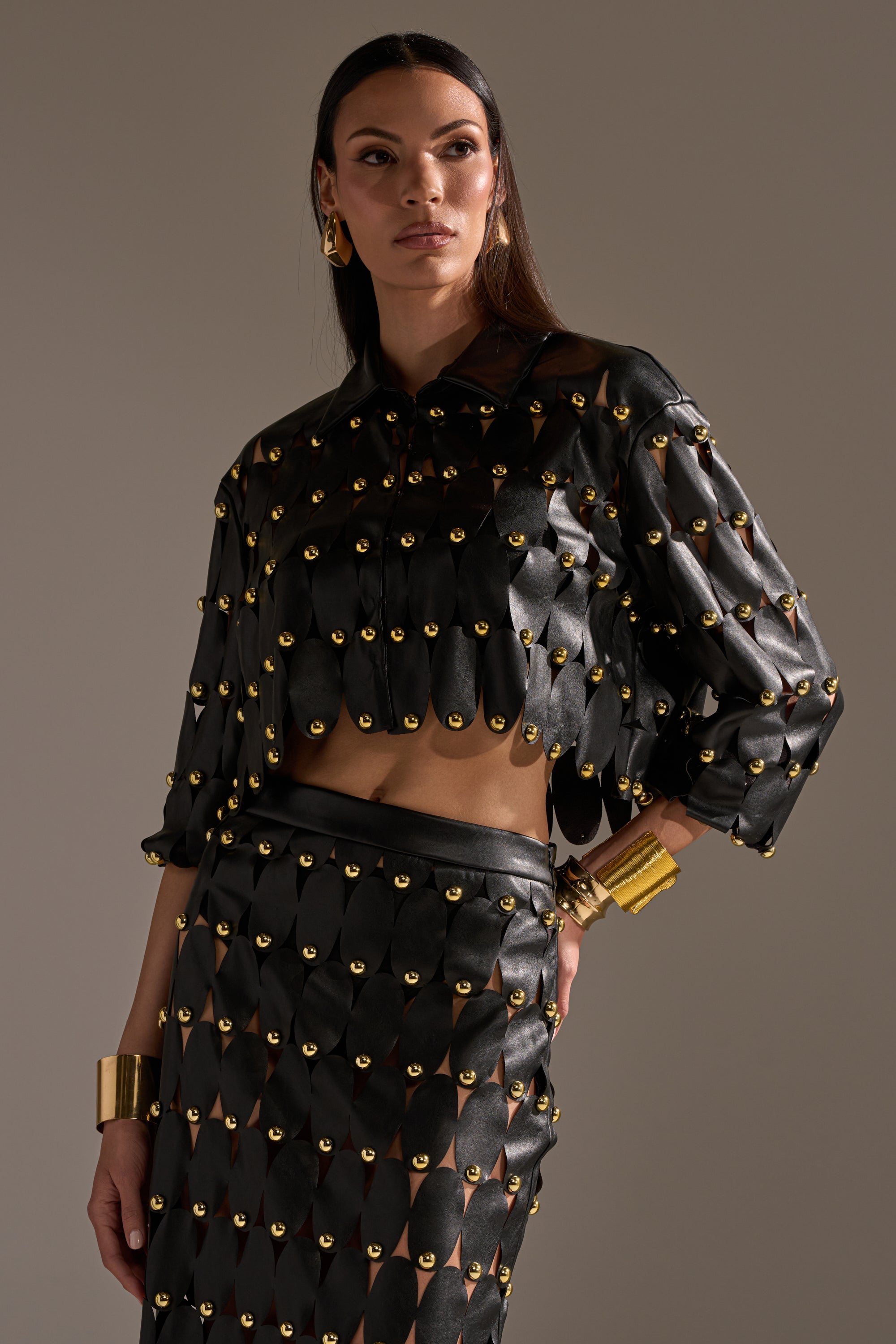 A woman models THE SNEAK PEEK STUDDED PU BLOUSE IN BLACK with a matching skirt, both featuring gold circular studs. She accessorizes with gold hoop earrings and chunky bracelets, posing against a neutral background. 7