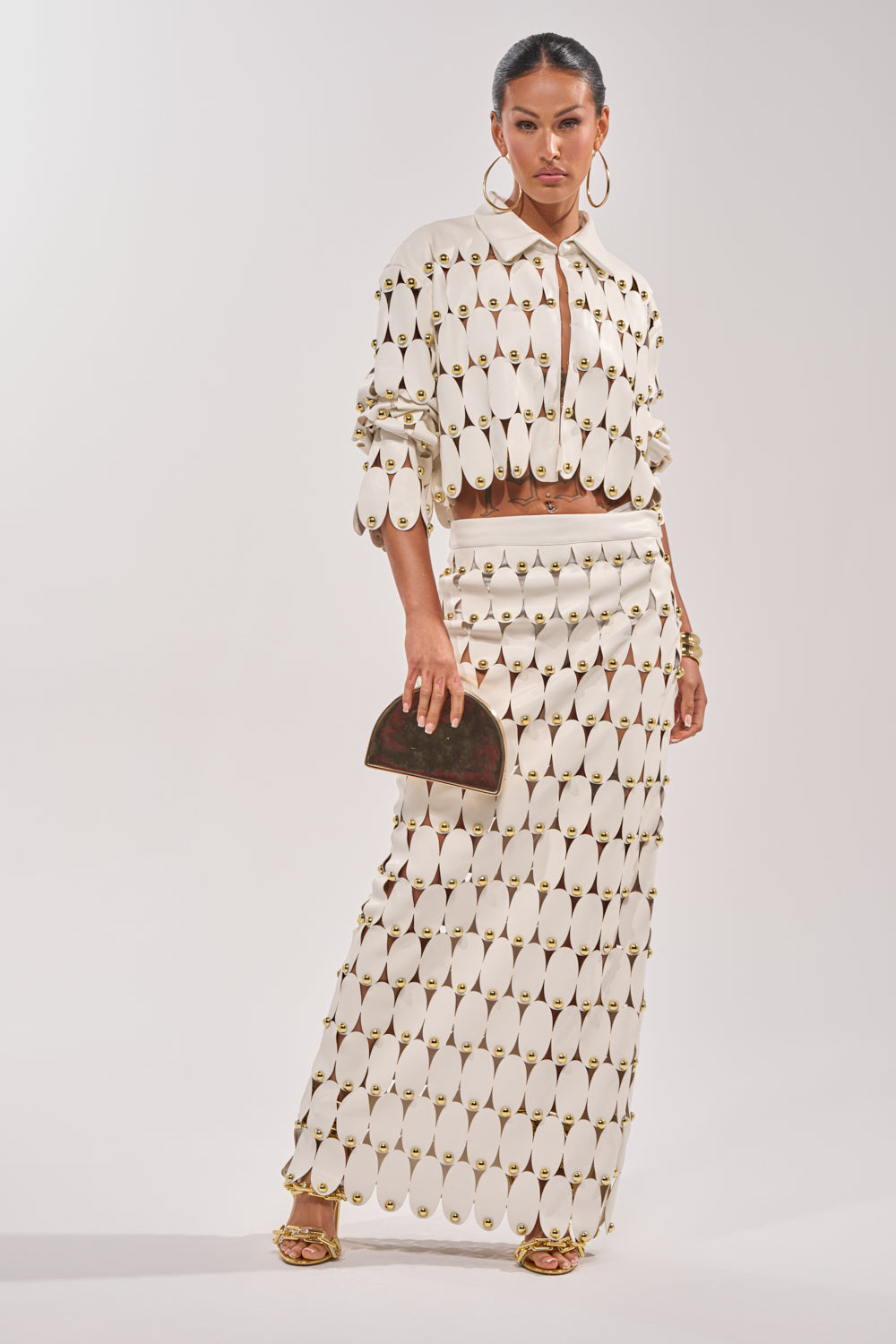 A woman poses in THE SNEAK PEEK STUDDED PU MIDI SKIRT IN WHITE with a matching cream cropped jacket, gold hoop earrings, gold heels, and a small brown clutch against a plain light background. 0