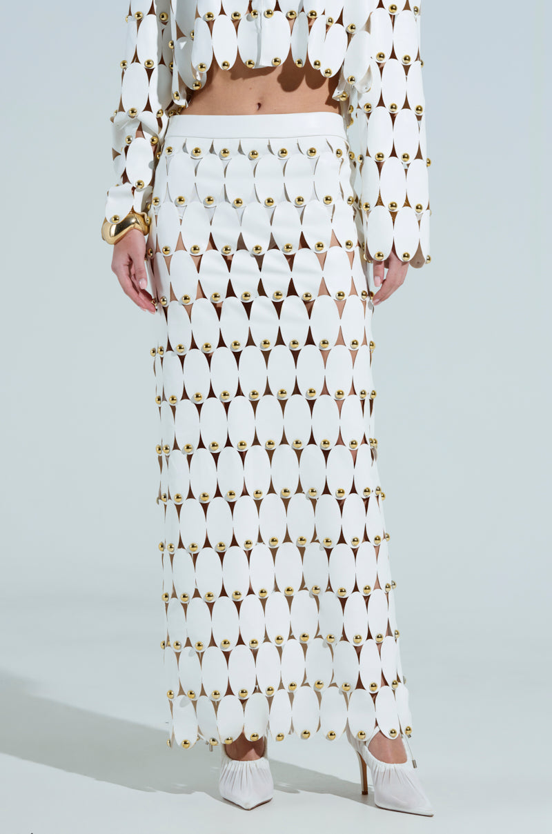 A person wears a geometric fashion ensemble with circular cutouts and brass accents, featuring THE SNEAK PEEK STUDDED PU MIDI SKIRT IN WHITE. The outfit is completed with studded white high-heeled shoes against a plain light gray background.