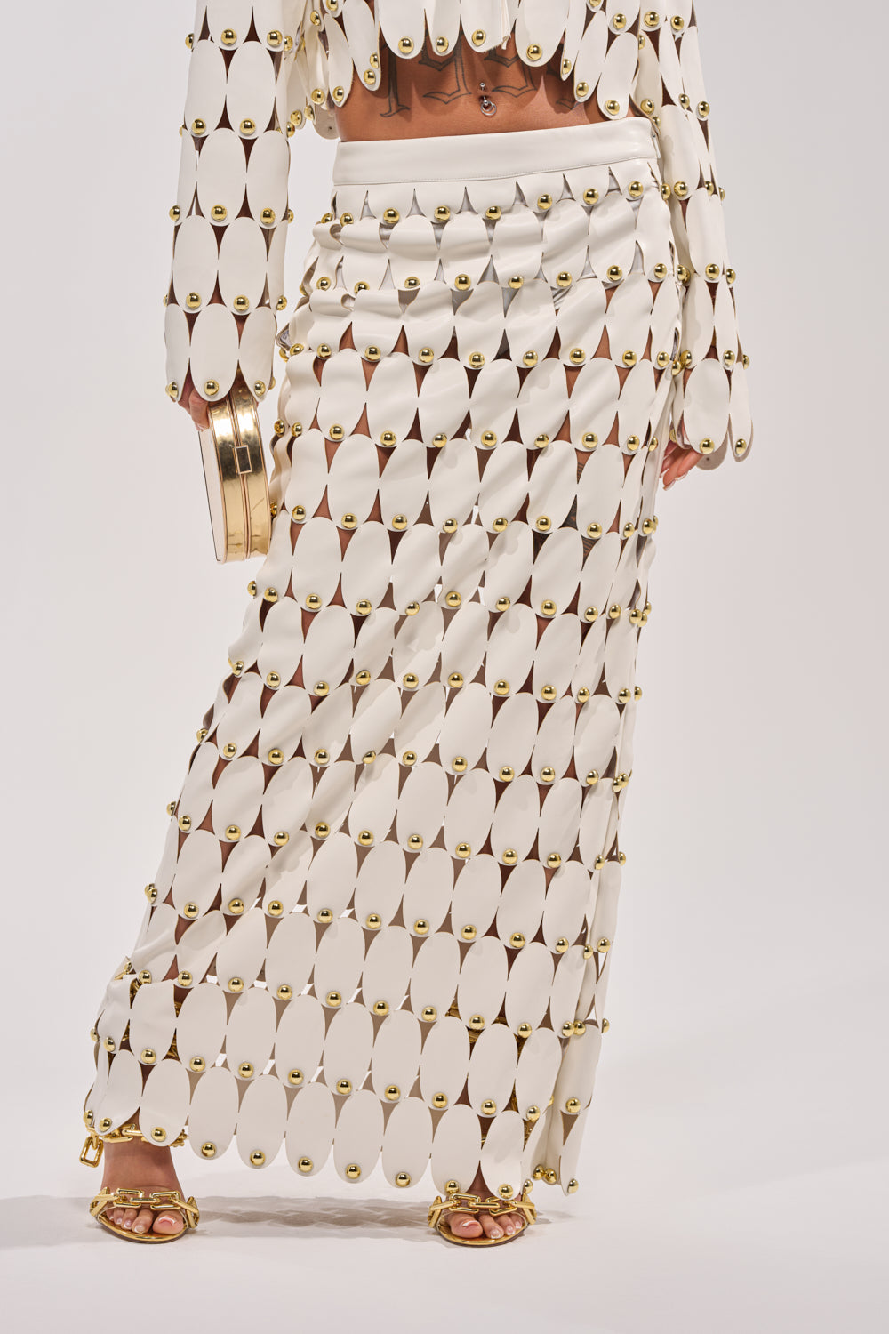 A person wears THE SNEAK PEEK STUDDED PU MIDI SKIRT IN WHITE, styled with a matching top, gold sandals, and a gold clutch. The image is cropped to focus on the lower body. 1