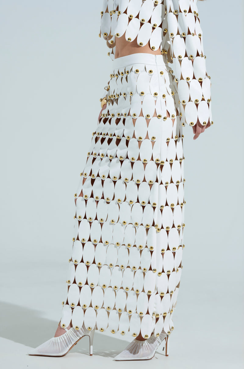 A person is wearing a white outfit made of oval faux leather pieces linked by gold rings, featuring THE SNEAK PEEK STUDDED PU MIDI SKIRT and a matching top. They are also in white high-heeled shoes against a neutral background.