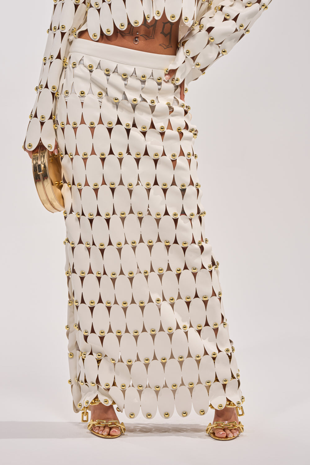 A person stands against a white background wearing THE SNEAK PEEK STUDDED PU MIDI SKIRT IN WHITE with a matching top featuring teardrop-shaped panels connected by gold rings, gold high-heeled sandals, and a round gold purse. 2