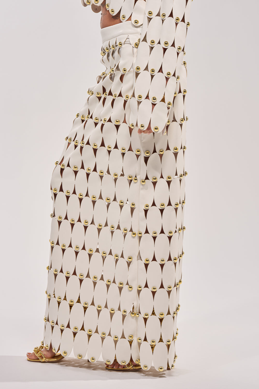 A person models THE SNEAK PEEK STUDDED PU MIDI SKIRT IN WHITE, featuring connected oval panels and gold fasteners, standing barefoot against a plain white background with only the lower body visible. 3
