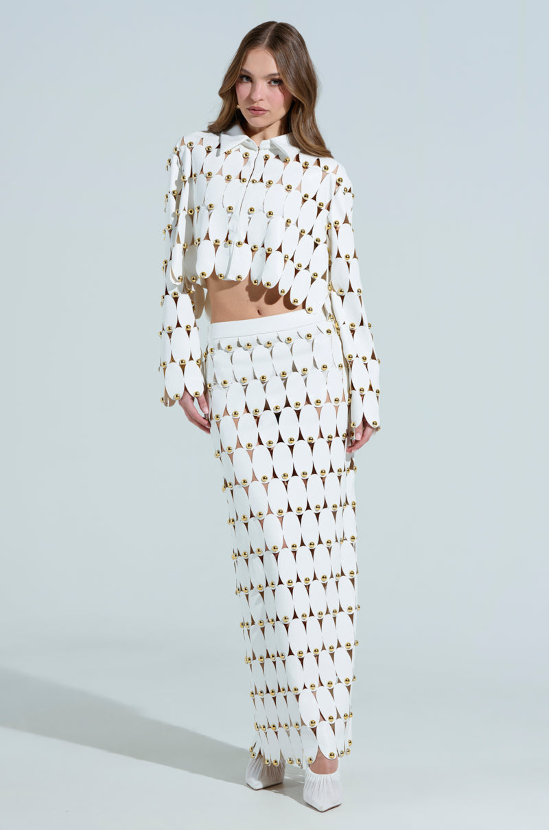 A woman wears a white long-sleeve top and THE SNEAK PEEK STUDDED PU MIDI SKIRT IN WHITE with a geometric cutout pattern. Her light brown hair complements the design, and her white shoes add a modern touch against the plain background.
