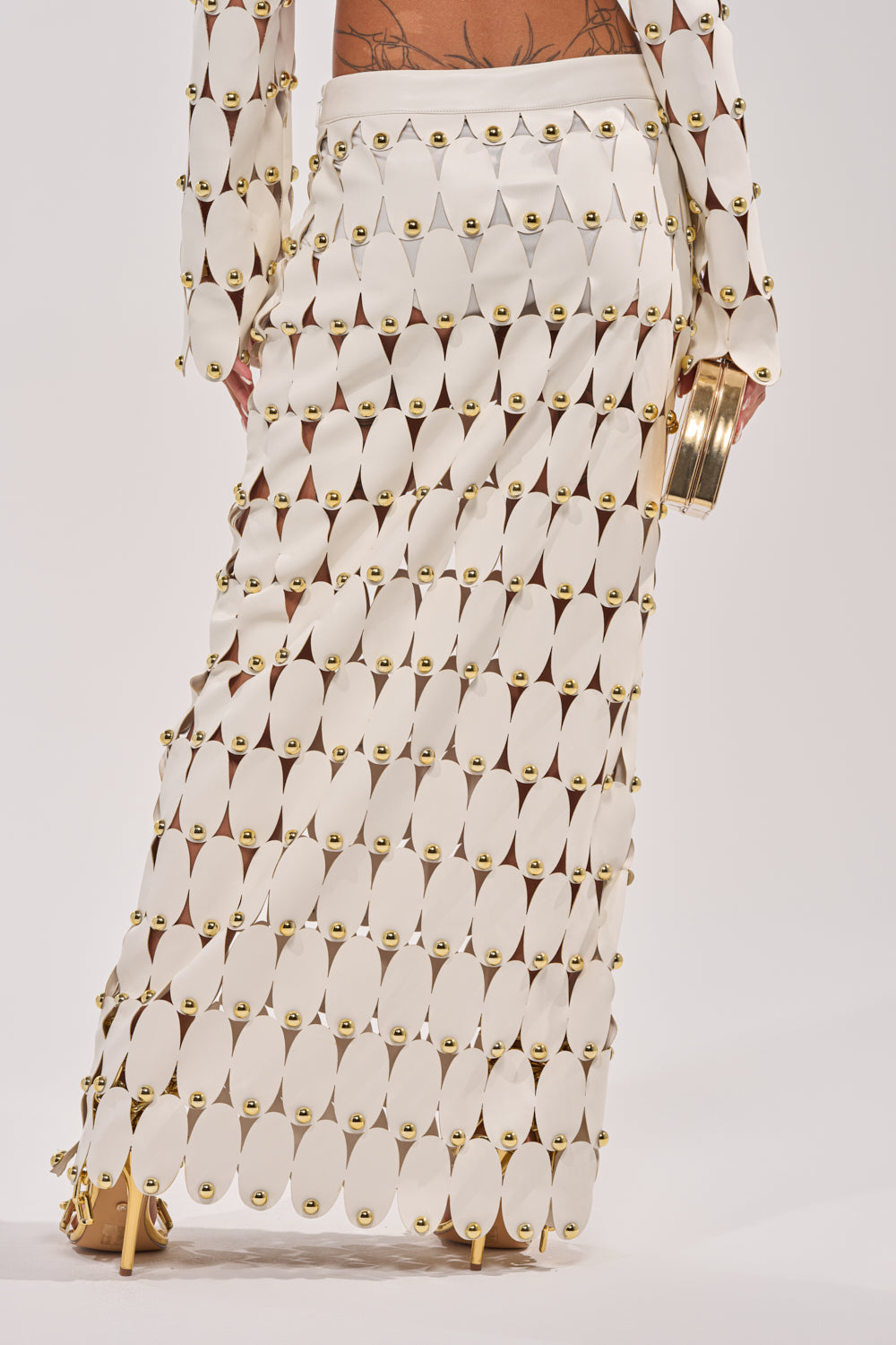 A person wears THE SNEAK PEEK STUDDED PU MIDI SKIRT IN WHITE—crafted from white faux leather panels joined by gold rings, featuring a geometric cutout design. The look is completed with a gold clutch against a plain white backdrop. 4