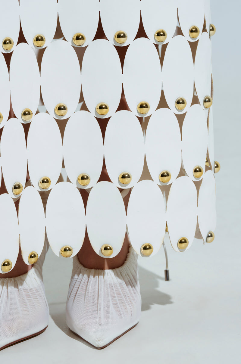 A detailed look at THE SNEAK PEEK STUDDED PU MIDI SKIRT IN WHITE, featuring large oval cutouts linked with gold studs, revealing hints of legs underneath. It's elegantly paired with white pointed-toe high-heeled shoes that boast gathered details.