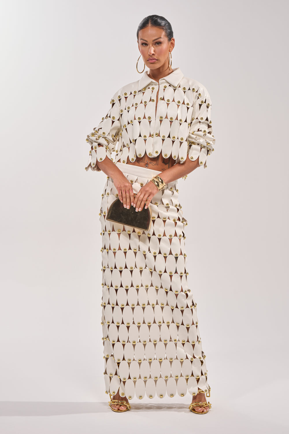 A woman poses against a plain backdrop wearing a cream long-sleeve top and THE SNEAK PEEK STUDDED PU MIDI SKIRT IN WHITE with oval cutouts and gold accents. She accessorizes with hoop earrings, strappy gold heels, and a brown clutch. 5