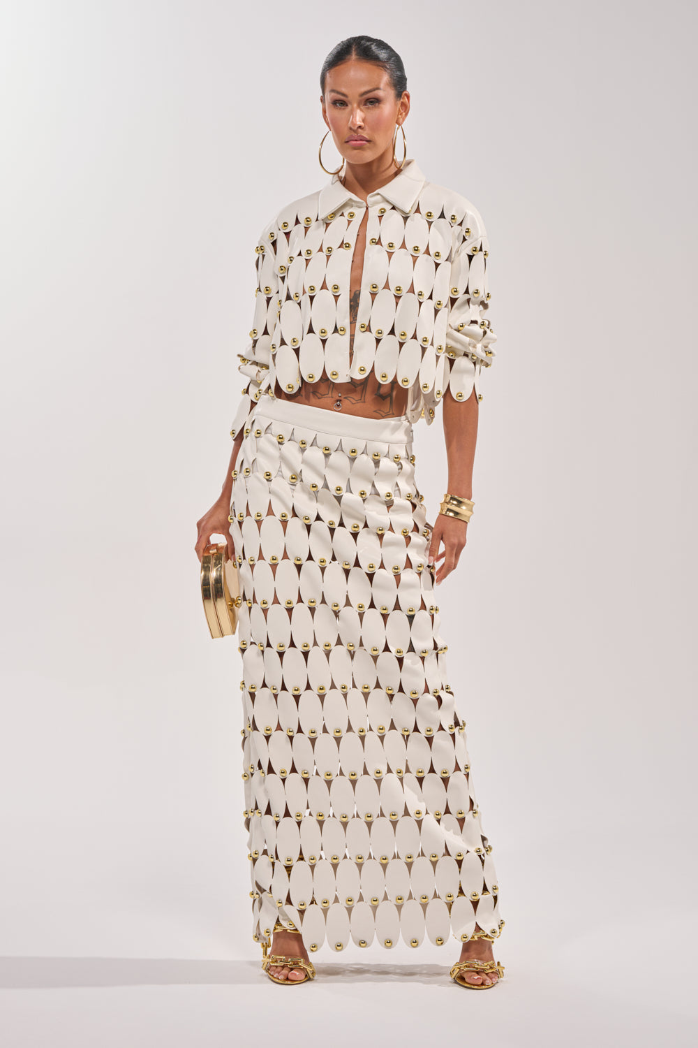A woman stands confidently in THE SNEAK PEEK STUDDED PU MIDI SKIRT IN WHITE paired with a cream geometric cut-out top, gold accents, large hoop earrings, gold bangles, and a round clutch. 6