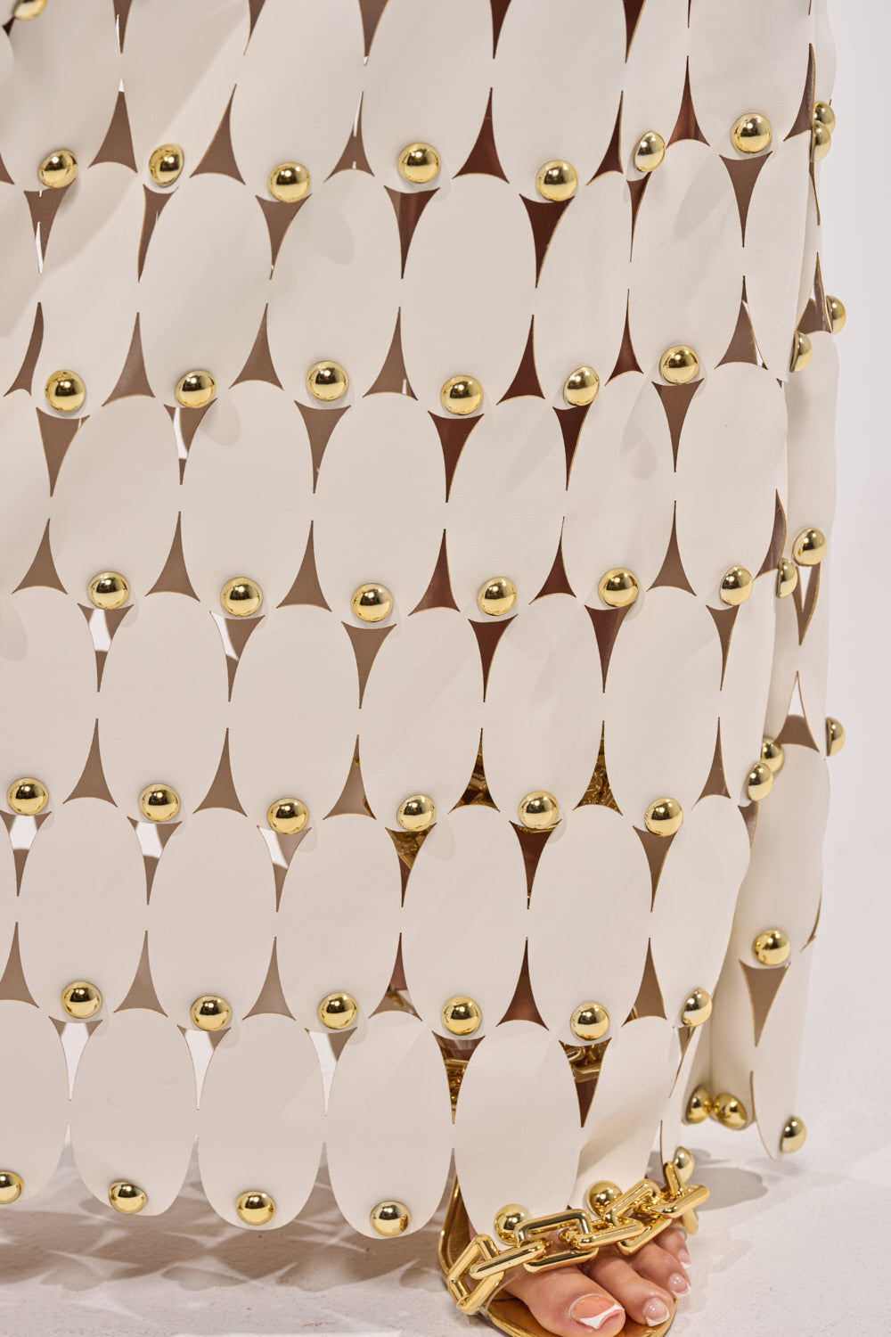 A close-up of THE SNEAK PEEK STUDDED PU MIDI SKIRT IN WHITE, featuring gold-studded oval panels joined by small gold spheres, with a gold chain sandal on a person’s foot at the bottom. 7