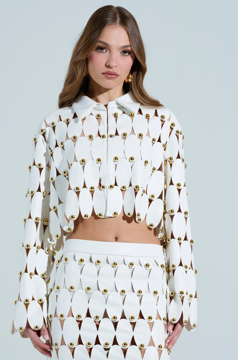 A woman wears the unique white SNEAK PEEK Studded PU Crop Cardigan with gold accents. The outfit has oval cutouts connected by gold fasteners, complemented by her long hair against a plain backdrop.