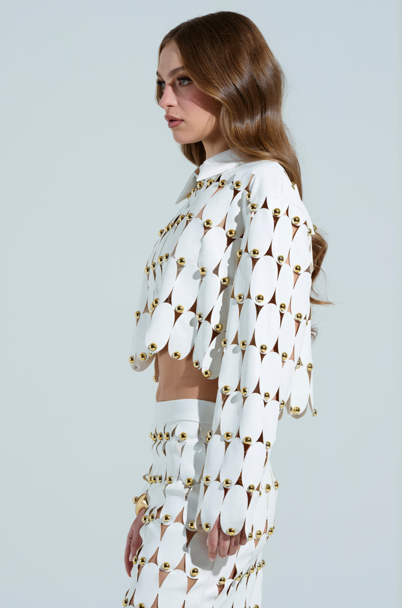 A person with long, wavy brown hair flaunts THE SNEAK PEEK STUDDED PU CROP CARDIGAN IN WHITE. The unique white outfit features oval cutouts with gold studs, scallop hemlines on a cropped top, and matching bottoms that form a striking geometric pattern against a light gray background.
