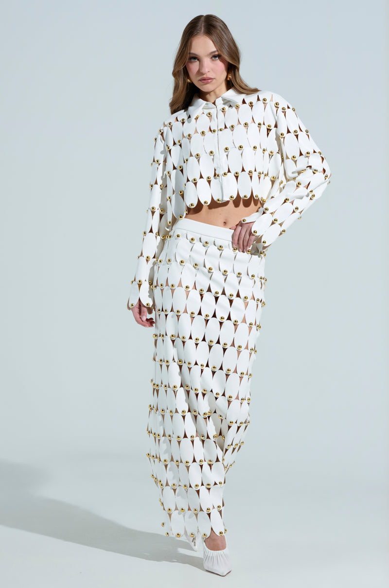 A person confidently poses in a matching white outfit featuring oval cutouts, scallop hemlines, and gold accents. They wear "The Sneak Peek Studded PU Crop Cardigan in White" with a long skirt and white pointed shoes, set against a light gray background.