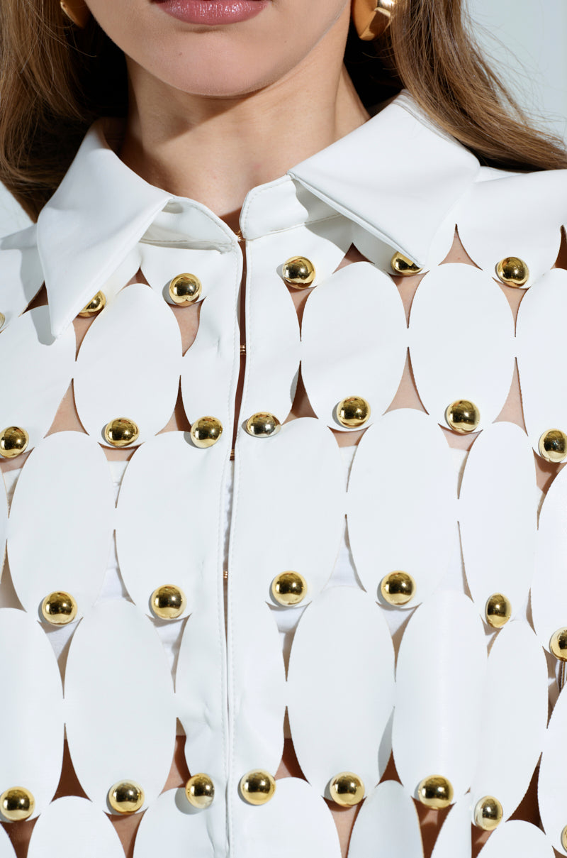 A close-up of someone in THE SNEAK PEEK STUDDED PU CROP CARDIGAN IN WHITE, featuring oval cutouts and gold studs. Scallop hemlines and a pointed collar enhance its geometric elegance. Long hair and gold hoop earrings complete the stylish look.
