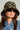 A woman with long, brown hair wears the PAINT ME CAMO BUCKET HAT featuring a painterly splatter design, accompanied by black sunglasses and a denim jacket. Her neutral expression is set against a plain background.