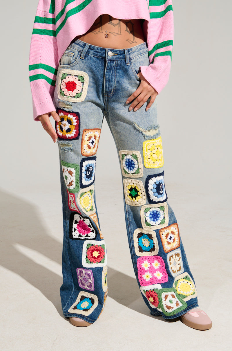 A person is wearing the PEACE & LOVE CROCHET EMBROIDERED MID RISE DENIM, which is adorned with colorful square patches. The denim pairs perfectly with a pink and green striped cropped sweater. The vibrant patches showcase floral and geometric designs, adding flair to the chic ensemble.