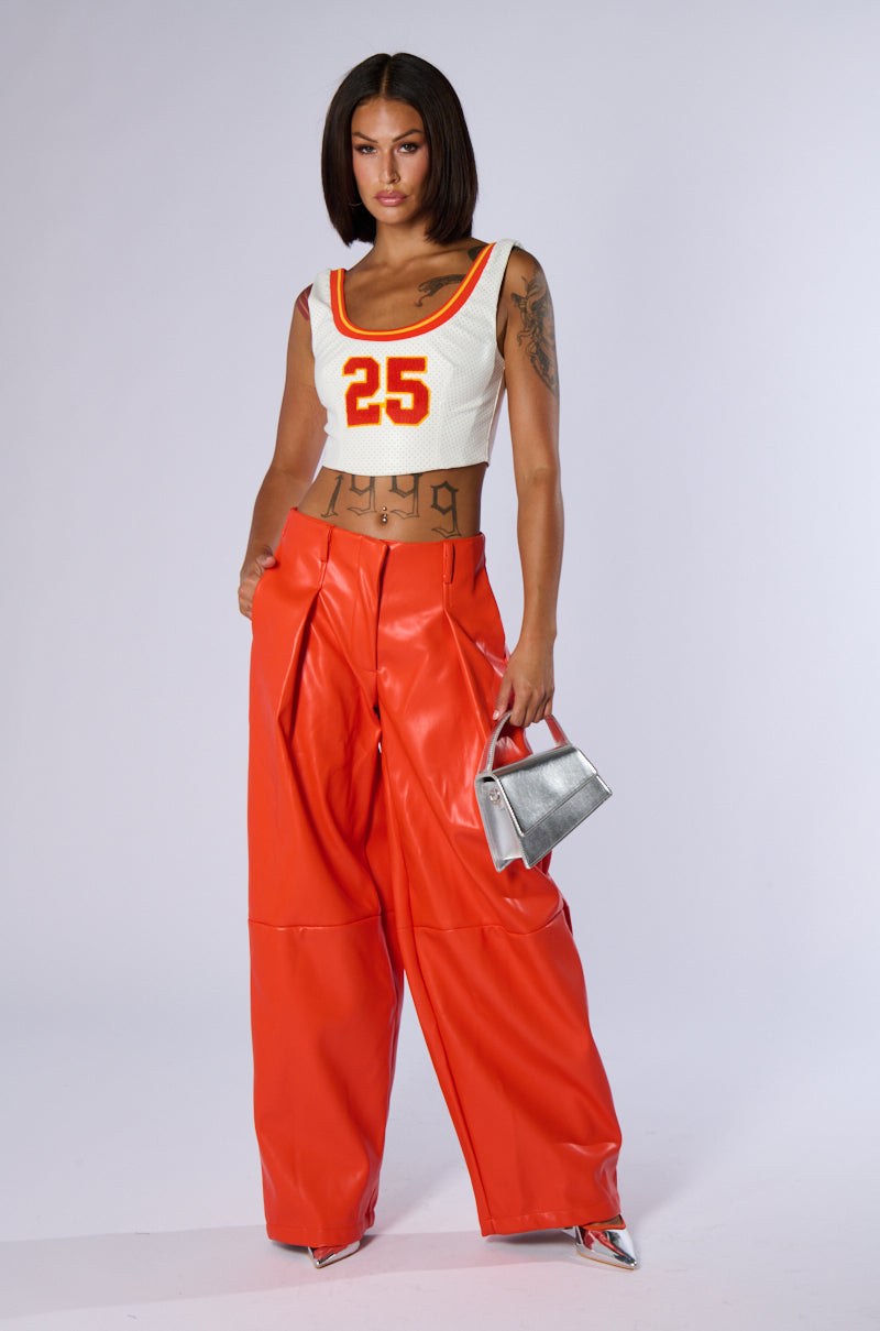 A person with shoulder-length dark hair stands confidently with one hand on their hip. They wear a white sleeveless crop top featuring the number "25" in red and orange, neon high-rise wide-leg faux leather BABY COME OVER PU PANT IN ORANGE, and hold a small silver handbag, paired with silver shoes.