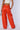 A person is standing with their back facing the camera, wearing high-waisted, wide-leg BABY COME OVER PU PANTS in orange from the AKIRA Label. They hold a small silver handbag in their left hand, and a portion of a white crop top is visible. The background is plain and light-colored.