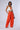 A person stands confidently against a plain background, wearing a white crop top featuring a red and orange "25" design and the BABY COME OVER PU PANT IN ORANGE from the AKIRA Label. They hold a small silver purse and wear heels, completing the fashionable ensemble.