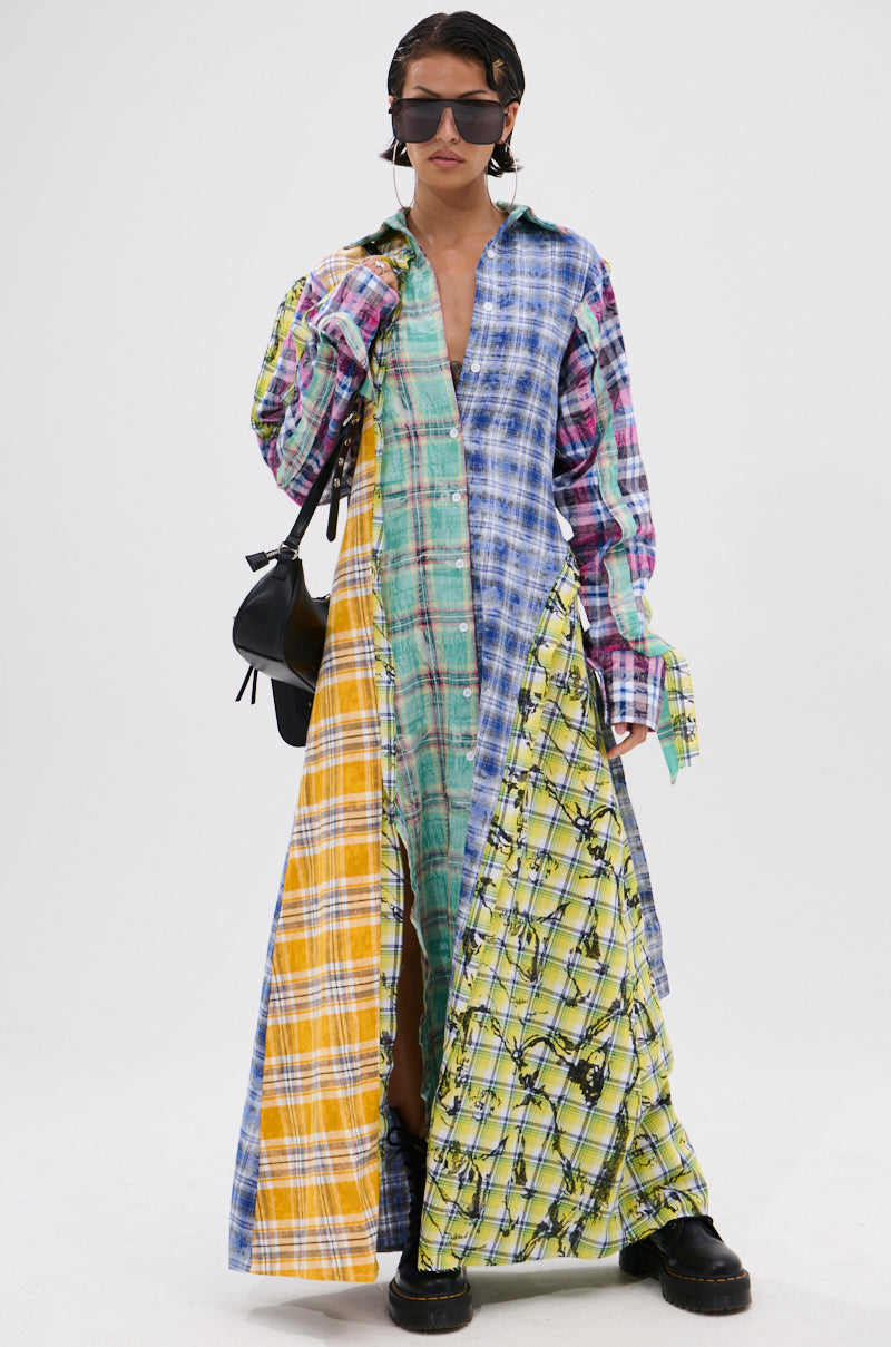 A person in the STAR OF THE SHOW PLAID MAXI DRESS with a patchwork design and self-tie waist stands against a plain background, accessorized with sunglasses, short dark hair, black boots, and a black handbag.