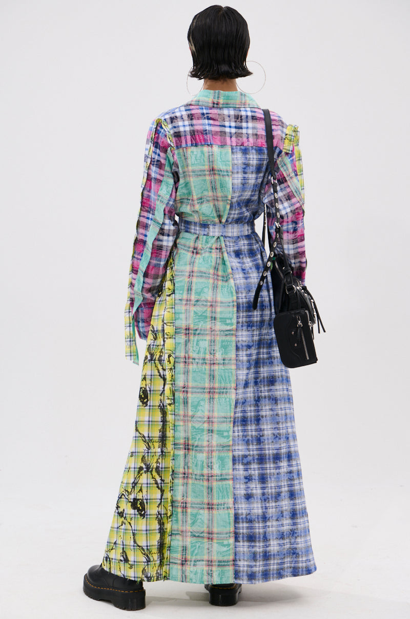 A person with short dark hair faces away, wearing the STAR OF THE SHOW PLAID MAXI DRESS featuring a patchwork design in pink, blue, green, and yellow. They carry a black handbag and wear black platform shoes against a plain white background.