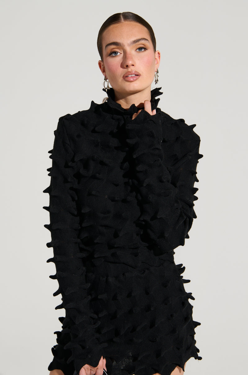 A woman with long brown hair is posing confidently in a unique black turtleneck dress near her chin, featuring the super stretchy ABSTRACT ART TEXTURED KNIT LONG SLEEVE TOP. The garment showcases an intricate, wavy 3D pattern and she gazes directly at the camera against a plain, light background. 0