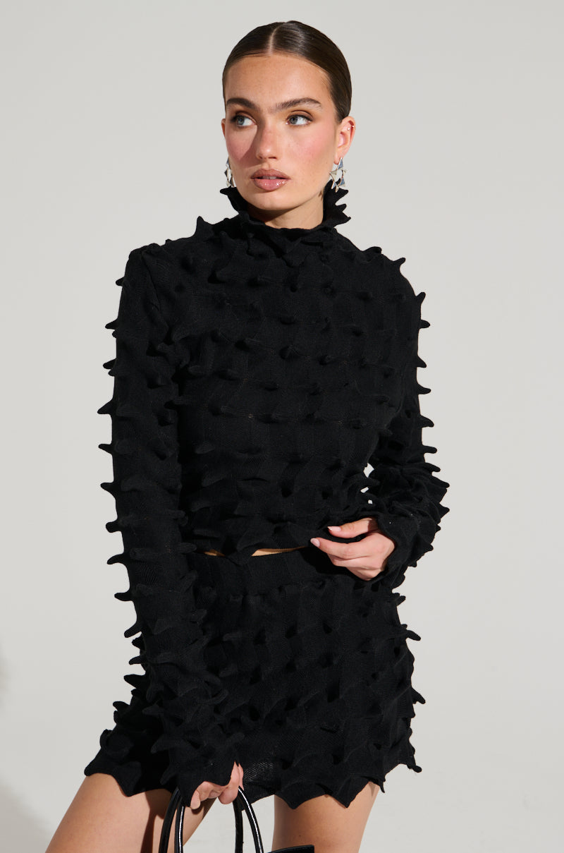 A woman dressed in an ABSTRACT ART TEXTURED KNIT LONG SLEEVE TOP and a matching skirt, both featuring a unique spiky pattern reminiscent of abstract art, stands against a plain background. Clutching a small black bag in one hand, she gazes thoughtfully to her right. 1