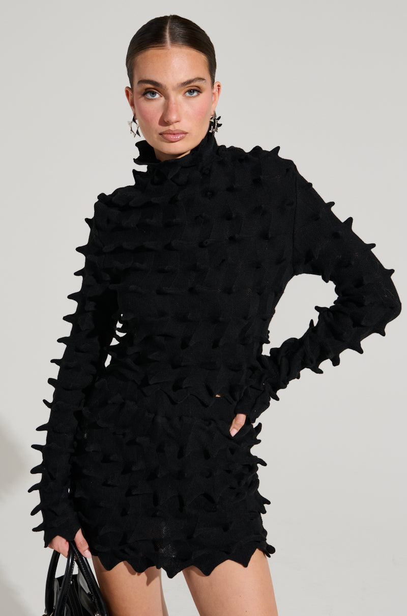 A woman stands confidently against a plain background, wearing an ABSTRACT ART TEXTURED KNIT LONG SLEEVE TOP with a high collar and long sleeves. She holds a black handbag and looks directly at the camera, her hair pulled back, and wears statement earrings. The top's fabric resembles abstract art textured knit, adding depth to her look. 2