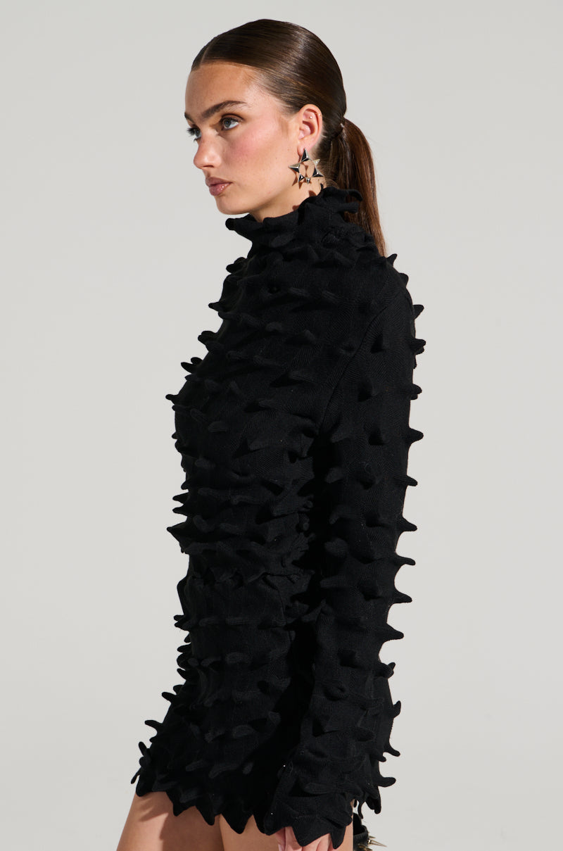A person with long brown hair tied back is wearing a distinctive black turtleneck top from our "Abstract Art Textured Knit Long Sleeve Top" collection, featuring an abstract art textured knit of raised, pointed details. They have star-shaped earrings and are turned slightly to the side, posing against a plain, light grey background. 3