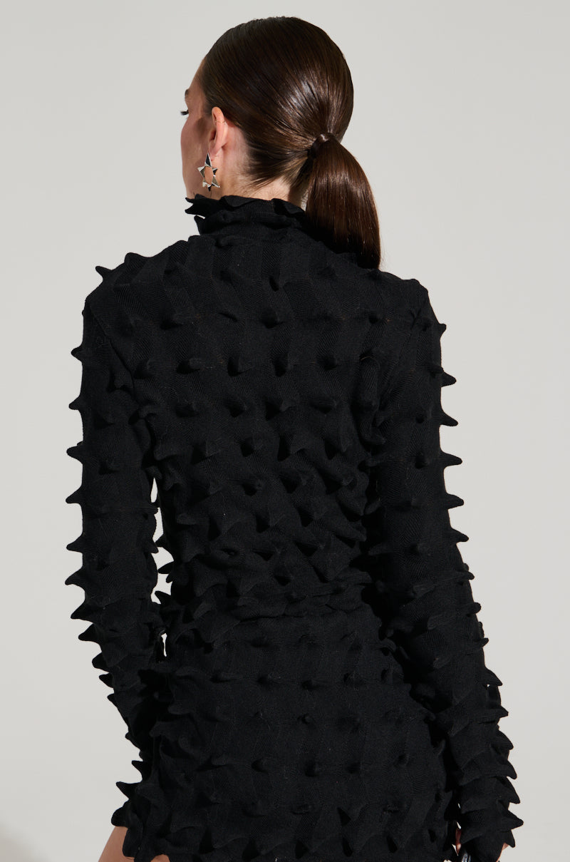 A person with long, dark hair is seen from the back, wearing the ABSTRACT ART TEXTURED KNIT LONG SLEEVE TOP. This black turtleneck top features a unique, raised abstract art pattern. Star-shaped earrings dangle elegantly against a plain, light-colored background. 4