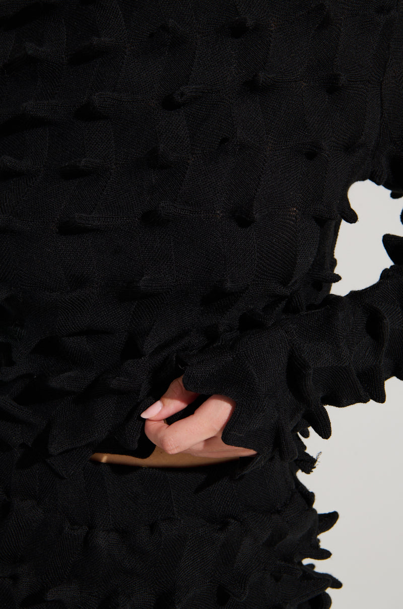 A close-up image showcases someone wearing the ABSTRACT ART TEXTURED KNIT LONG SLEEVE TOP, a black sweater featuring raised, spiky patterns reminiscent of abstract art. Their manicured hand peeks through the super stretchy fabric at the waist, while the black turtleneck covers most of their upper body, effortlessly highlighting its distinctive 3D design. 7