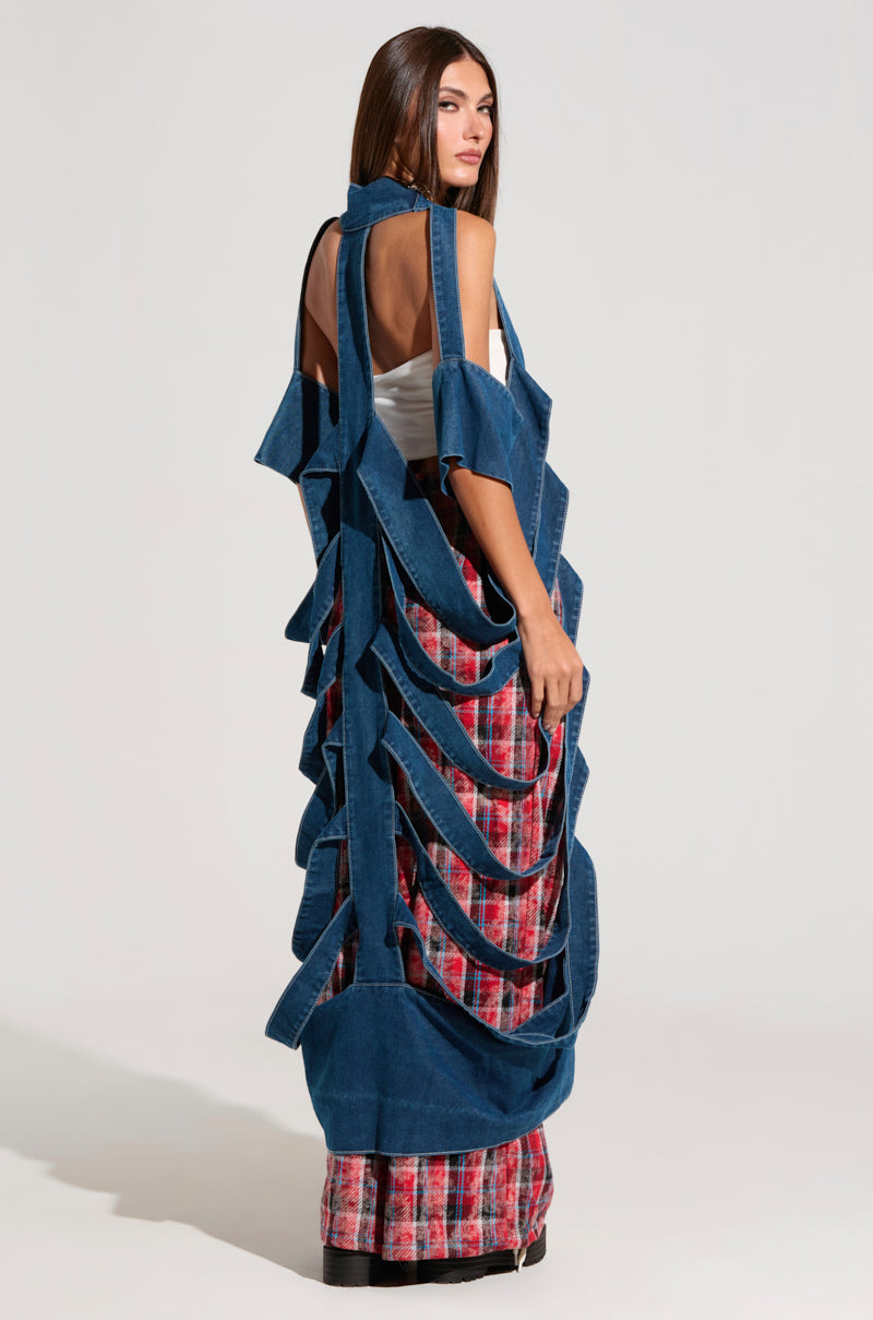A woman with long brown hair, dressed in a white top and red plaid skirt, completes her outfit with a GEO CUTOUT DENIM DUSTER characterized by multiple straps. She stands facing backward, glancing over her shoulder with a neutral expression in the high-fashion ambiance of the white studio.