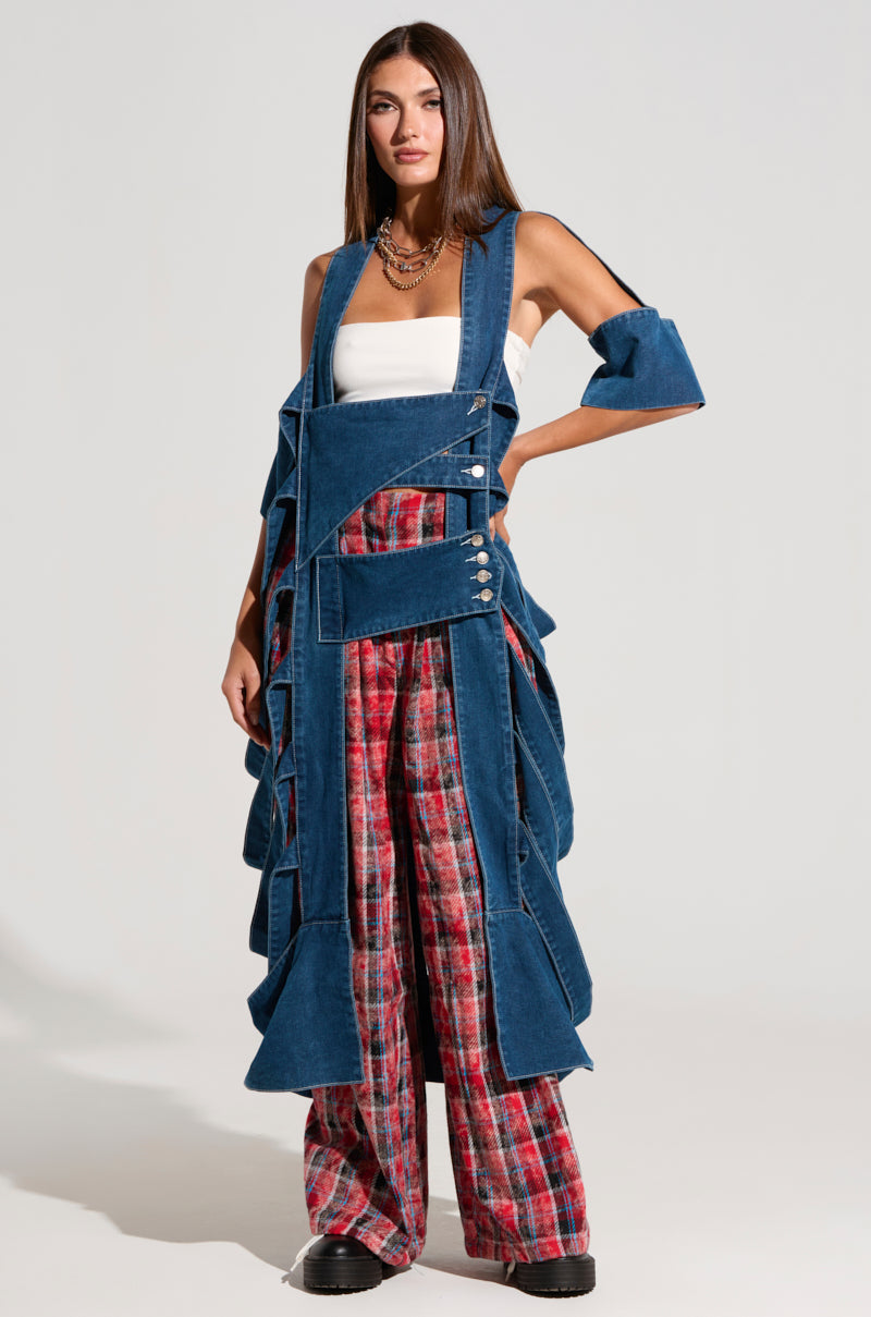 A woman with long brown hair stands confidently in the GEO CUTOUT DENIM DUSTER over a white tube top and red plaid wide-leg pants. She accessorizes with multiple necklaces and black platform shoes, posing against a plain white background, showcasing her high-fashion finish.