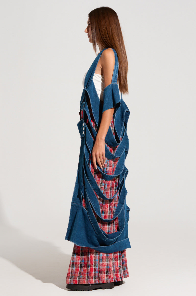A woman with long brown hair stands sideways, showcasing a high-fashion look that includes a white top and the GEO CUTOUT DENIM DUSTER styled as a vest. She complements this distinctive jacket with red plaid pants that flare at the bottom, all set against a plain, light-colored background.