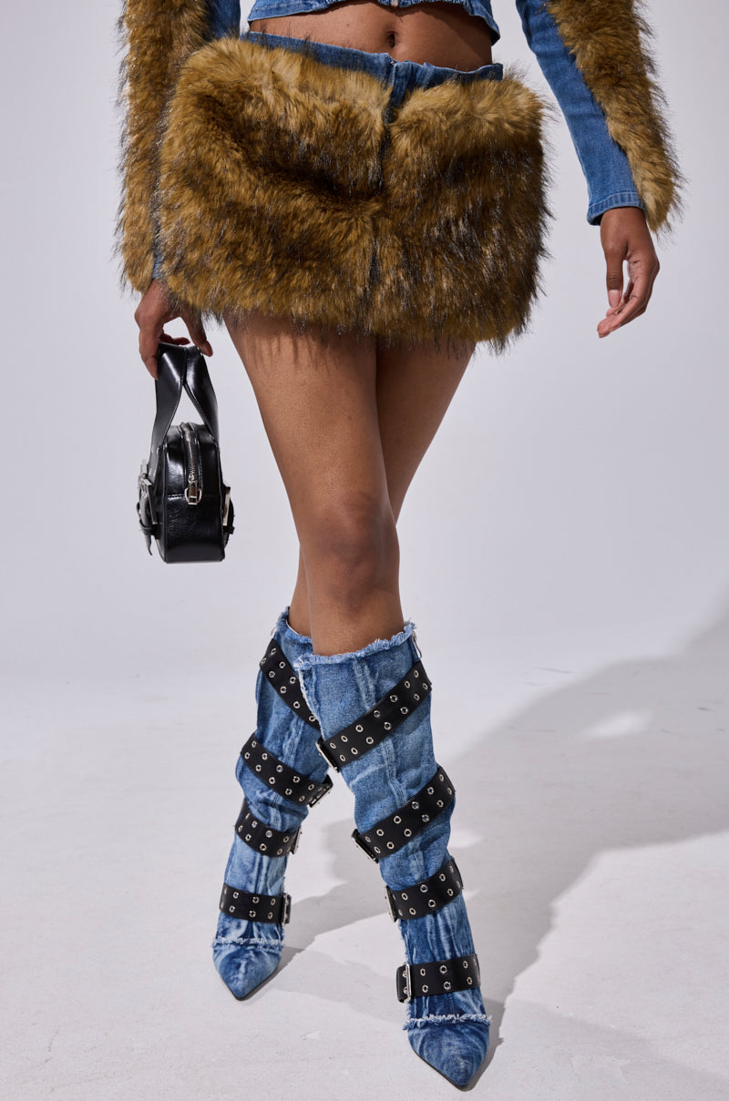 A person wearing the OUT OF THE BLUE FUR MINI SKIRT, paired with denim boots featuring black buckles and eyelets, and holding a small black handbag. Only the waist down is visible against a plain background.