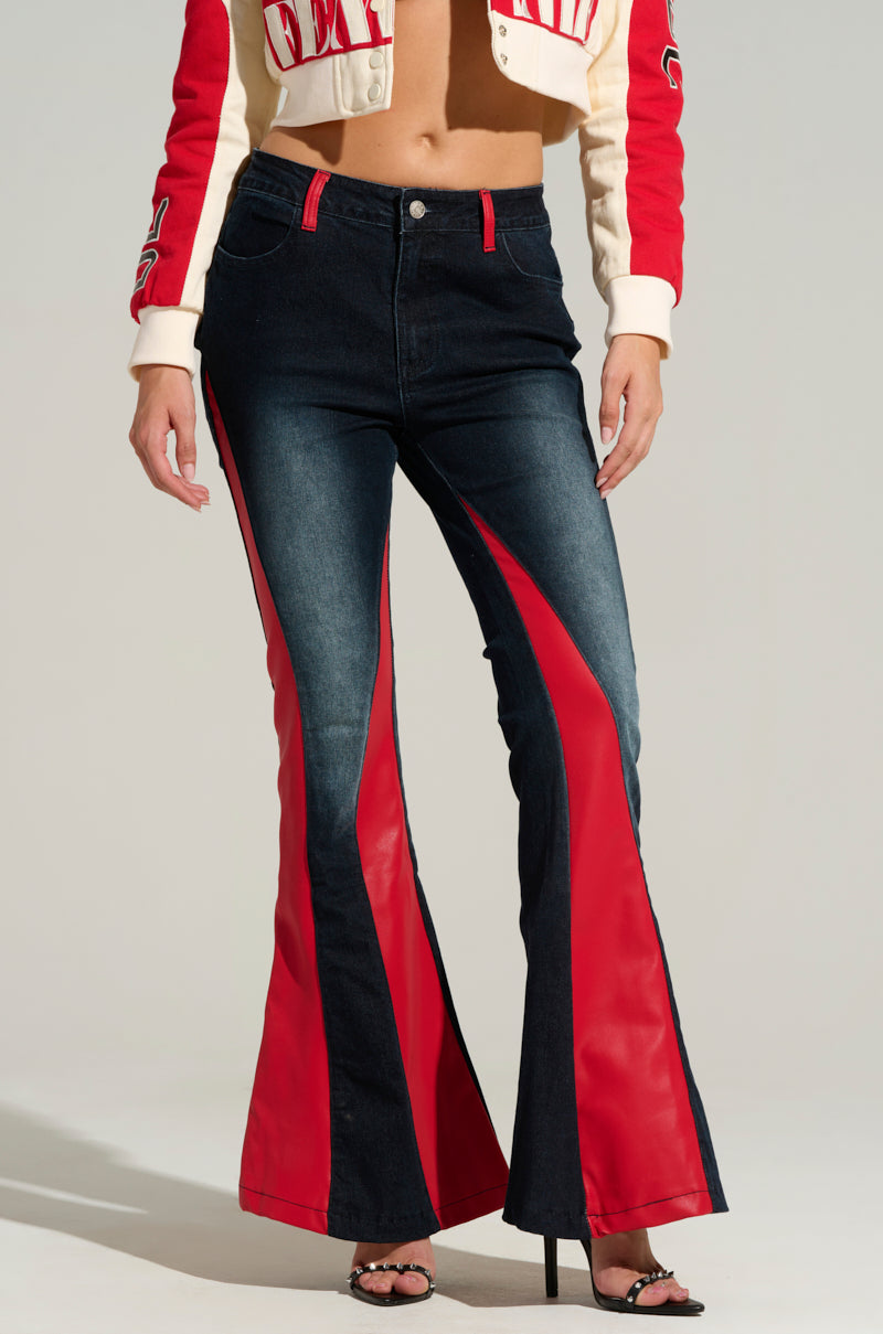 A person is sporting the STELLA BELLA FLARE DENIM PANT, featuring striking red panels extending from the thighs to the ankles. Additionally, they have paired it with a high-rise, cropped jacket in shades of red and cream, along with black heeled sandals. The photograph is captured in a studio environment.