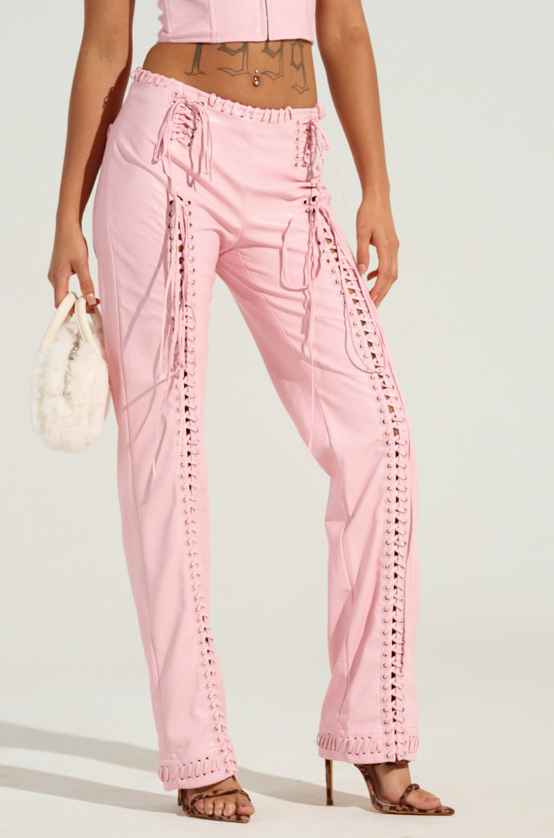 A person wearing the FLASHING LIGHTS FAUX LEATHER PANT in light pink and a matching crop top holds a small white fuzzy handbag. Their feet are adorned with brown open-toed heels, revealing a tattoo on their midsection. The background is plain and light-colored.