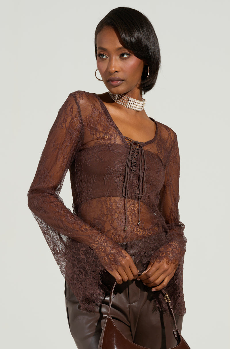 Dressed in the Starlight Long Sleeve Lace Blouse in Brown, featuring flared sleeves and a delicate lace-up front, the individual poses against a simple backdrop. The sheer lace design brings an elegant touch to their ensemble, accentuated by hoop earrings and a choker necklace. They hold a dark purse with both hands.
