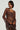 Dressed in the Starlight Long Sleeve Lace Blouse in Brown, featuring flared sleeves and a delicate lace-up front, the individual poses against a simple backdrop. The sheer lace design brings an elegant touch to their ensemble, accentuated by hoop earrings and a choker necklace. They hold a dark purse with both hands.