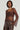 The individual is wearing the Starlight Long Sleeve Lace Blouse in brown, which features a sheer lace design, paired elegantly with brown leather pants. A pearl necklace and hoop earrings enhance the sophistication, while their hand rests casually in their pocket against a simple light background.