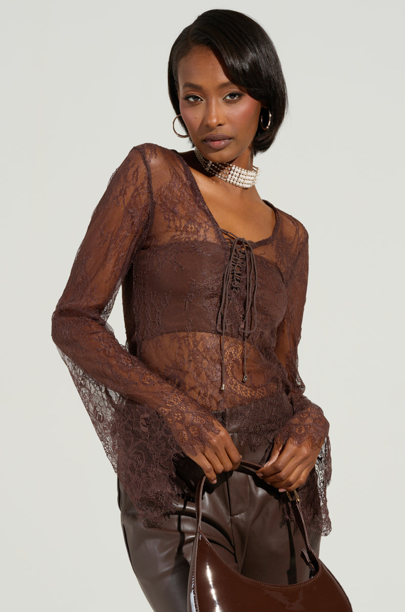 A person wearing the STARLIGHT LONG SLEEVE LACE BLOUSE IN BROWN, which features a sheer fabric and a subtle floral pattern, is holding a brown handbag. They have a sleek hairstyle and are accessorized with hoop earrings and a choker necklace, set against a plain background.