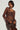 A person wearing the STARLIGHT LONG SLEEVE LACE BLOUSE IN BROWN, which features a sheer fabric and a subtle floral pattern, is holding a brown handbag. They have a sleek hairstyle and are accessorized with hoop earrings and a choker necklace, set against a plain background.