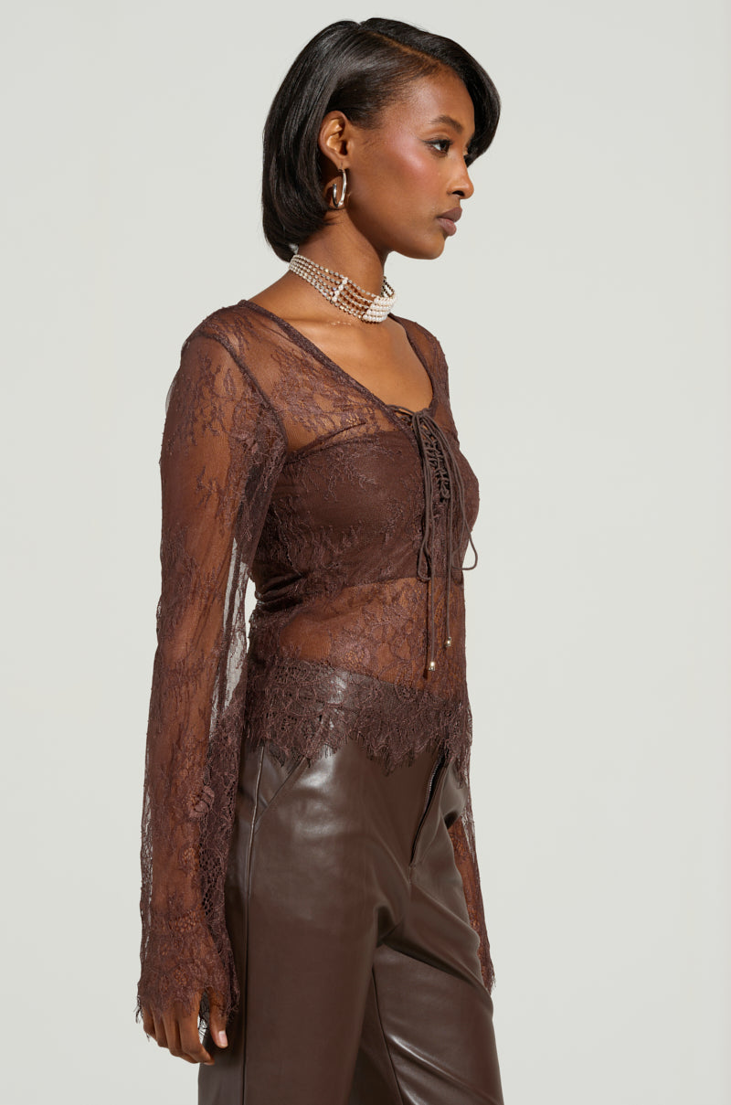 A person wearing the Starlight Long Sleeve Lace Blouse in brown, featuring sheer lace fabrication and a floral pattern with a lace-up front, paired with dark leather pants. They have short, styled hair and are accessorized with a choker necklace and earrings, set against a plain background.
