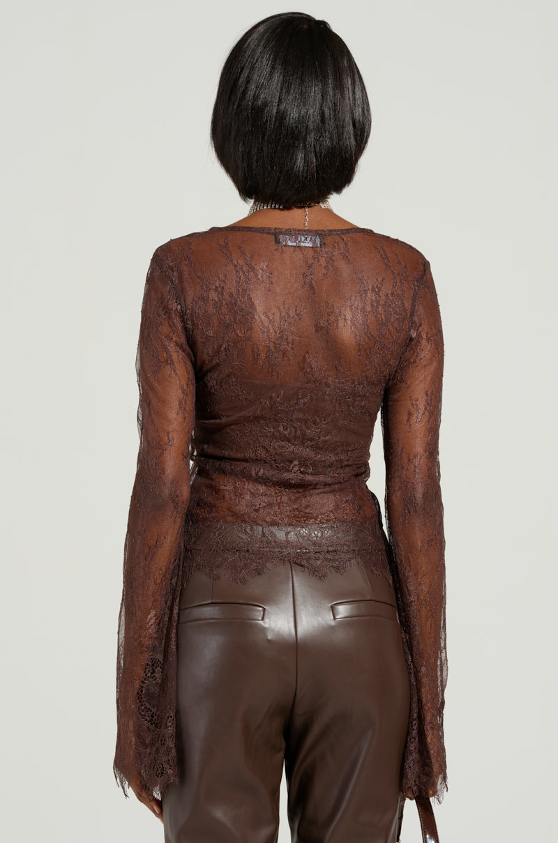 A person with short dark hair is seen from behind, wearing the Starlight Long Sleeve Lace Blouse in Brown, featuring a delicate floral pattern. They pair it with brown leather pants against a plain, light-colored background that accentuates the elegant design of their attire.