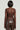 A person with short dark hair is seen from behind, wearing the Starlight Long Sleeve Lace Blouse in Brown, featuring a delicate floral pattern. They pair it with brown leather pants against a plain, light-colored background that accentuates the elegant design of their attire.