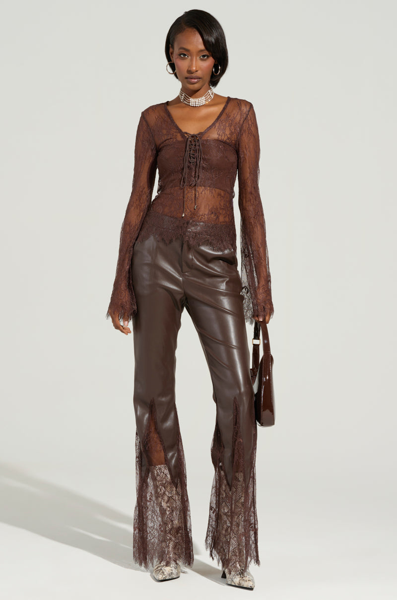A person stands against a plain background, wearing the STARLIGHT Long Sleeve Lace Blouse in Brown paired with matching leather pants featuring sheer lace fabrication accents. They hold a small handbag and wear a choker necklace.