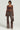 A person stands against a plain background, wearing the STARLIGHT Long Sleeve Lace Blouse in Brown paired with matching leather pants featuring sheer lace fabrication accents. They hold a small handbag and wear a choker necklace.