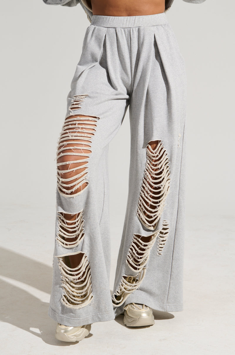 A person is wearing the STRESS HIM OUT SWEATPANT IN GREY, featuring a high-rise wide-leg fit with significant horizontal rips across the thighs and knees that reveal glimpses of their legs. They have paired the outfit with light-colored sneakers against a plain, light-colored background.