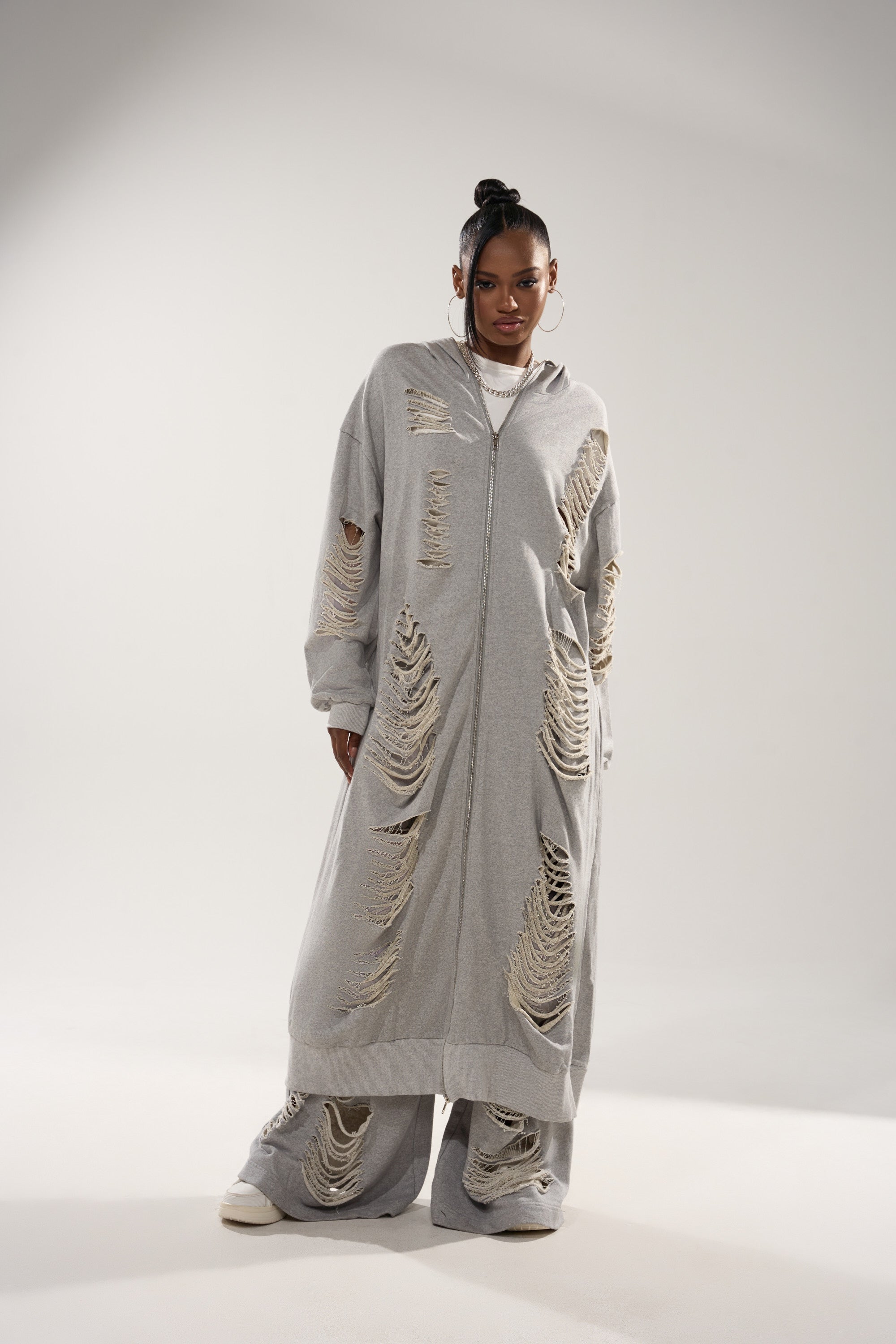 A woman stands in a studio wearing the STRESS HIM OUT LONGLINE SWEATSHIRT and matching distressed wide-leg pants with ripped details, accessorized with hoop earrings and white sneakers, against a plain white background. 1